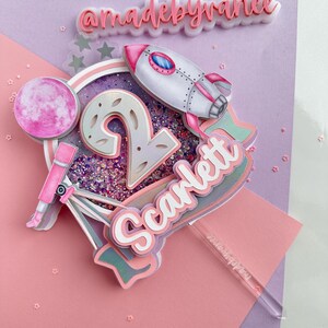 May include: A pink and purple cake topper featuring a rocket, a moon, a telescope, and the number 2. The name "Scarlett" is written on a pink and white banner. The text "@madebyvanee" is on a white background.