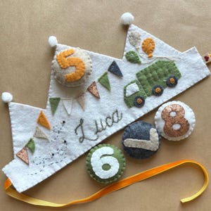 May include: A felt crown with a green truck, orange balloons, and a name Luca embroidered on it. The crown is decorated with orange, green, and white felt flags. There are also four felt circles with the numbers 5, 6, 7, and 8 on them.