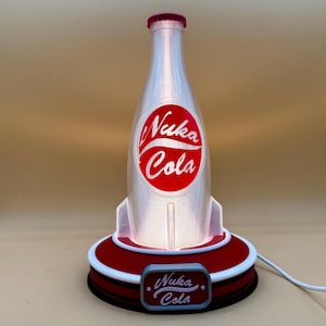 May include: A Nuka Cola bottle-shaped lamp with a red cap and a red and white logo. The lamp has a red base with a white trim and a small rectangular sign that says "Nuka Cola". The lamp is lit from within.