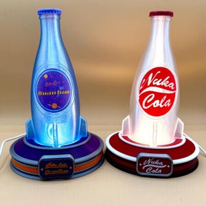 May include: Two bottle-shaped lamps with rocket ship bases. One is blue with "Nuka Cola Quantum" text, the other is white with "Nuka Cola" text. Both have a base with a small rectangular label with the same text. The lamps are lit.