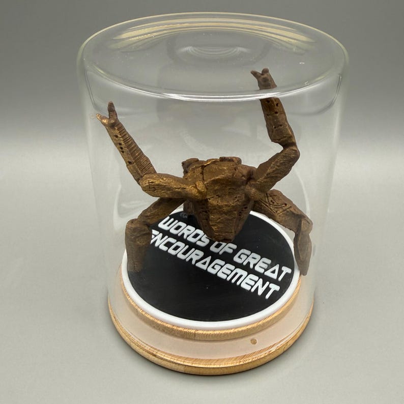 May include: A bronze-colored crab figurine with textured legs positioned inside a clear glass cloche. The crab rests on a circular black base featuring the white text "WORDS OF GREAT ENCOURAGEMENT" set upon a light-colored wooden circular stand.