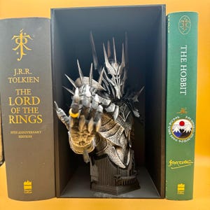 May include: A detailed bust of a character from The Lord of the Rings, with a hand extended, wearing a gold ring. The bust is displayed between two books, "The Lord of the Rings" and "The Hobbit". The bust is dark gray and silver.