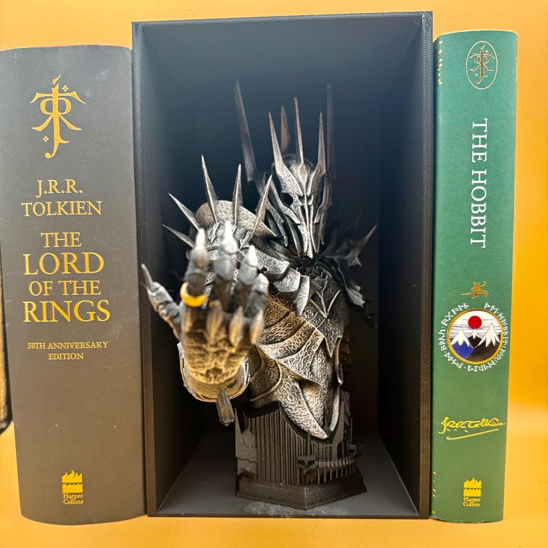 May include: A detailed bust of a character from The Lord of the Rings, with a hand extended, wearing a gold ring. The bust is displayed between two books, "The Lord of the Rings" and "The Hobbit". The bust is dark gray and silver.