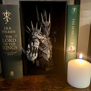 May include: A framed sculpture of a character from The Lord of the Rings, with a hand wearing a ring. Flanking the sculpture are two books: "The Lord of the Rings" and "The Hobbit." A lit white candle sits in front of the books, casting a warm glow.