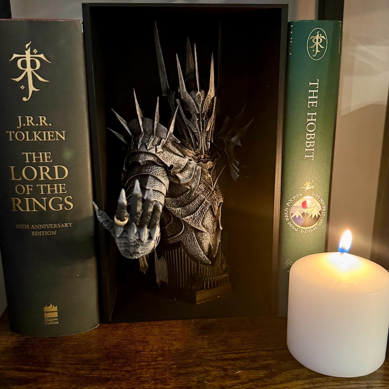 May include: A framed sculpture of a character from The Lord of the Rings, with a hand wearing a ring. Flanking the sculpture are two books: "The Lord of the Rings" and "The Hobbit." A lit white candle sits in front of the books, casting a warm glow.