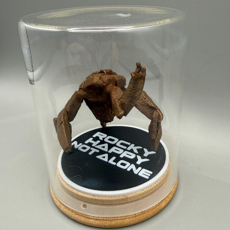 May include: A brown, textured, multi-limbed creature figurine enclosed in a clear glass dome. The figure stands on a circular black base with white text that reads ROCKY HAPPY NOT ALONE, set upon a light-colored wooden circular platform.