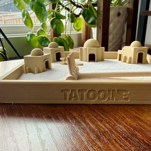 Könnte beinhalten: Ein Miniatur-Zen-Garten mit beigefarbenen Gebäuden, einem Rechen und weißem Sand. Die Holzbasis ist mit dem Wort "TATOOINE" graviert. Die Gebäude haben abgerundete Kuppeln und gewölbte Türen. Der Garten steht auf einer Holzoberfläche.