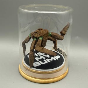 May include: A brown, multi-legged creature figurine with green accents, enclosed in a clear glass dome on a wooden base. The creature stands on a black circular platform featuring the white text "DIS MY BUMP" in bold, capital letters.