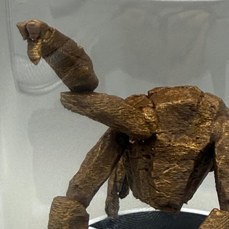 May include: A bronze-colored, textured figurine of a creature with a segmented, raised tail and a blocky, angular body structure. The surface displays a layered, 3D-printed appearance with visible horizontal lines across the metallic brown form.