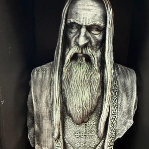 May include: A detailed bust of a wise, bearded figure with long hair and a flowing beard. The sculpture is in shades of gray and silver, with intricate detailing on the clothing and facial features. The bust is set against a dark background.