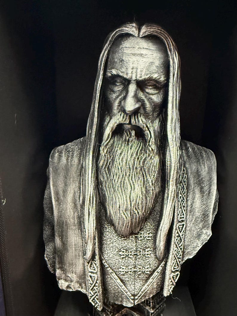 May include: A detailed bust of a wise, bearded figure with long hair and a flowing beard. The sculpture is in shades of gray and silver, with intricate detailing on the clothing and facial features. The bust is set against a dark background.