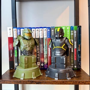 May include: Two detailed busts of video game characters, one green and one black, sit on a shelf. The green bust has a gold visor, while the black bust has yellow accents. Behind them are several video game cases for PS5 and Xbox consoles.