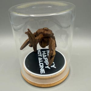 May include: A brown, textured, multi-legged creature figurine enclosed in a clear glass dome. The figure stands on a circular black base with white text that reads "ROCKY HAPPY NOT ALONE" set upon a light-colored wooden circular platform.