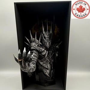 May include: A dark, armoured bust of a figure with a spiked helmet and gauntlet, featuring a gold-coloured ring on one finger. The figure is displayed within a black rectangular frame, with a circular 'MADE IN CANADA' logo in the top corner.