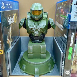 May include: A green and gray Master Chief bust, a character from the Halo video game series, is displayed on a pedestal. The bust is flanked by two video game cases, one for PS4 and one for PS5. The bust is made of a 3D printed material.