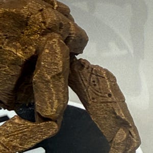 May include: A bronze-colored, textured figurine with a rough, rock-like surface. The object features a prominent limb extending outward, displaying a distinct geometric pattern etched into its side, consisting of a diamond shape with small dots.