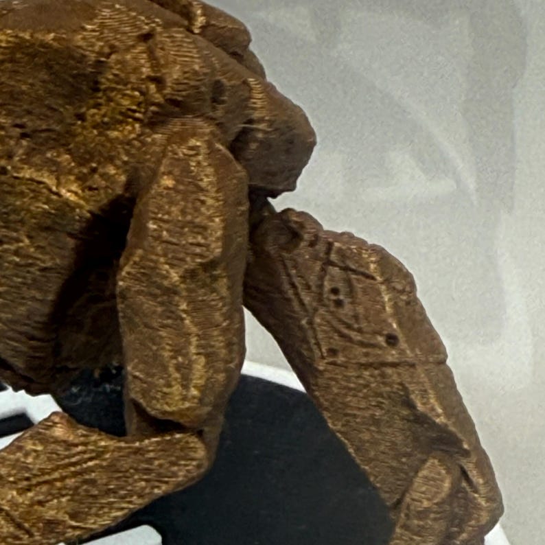 May include: A bronze-colored, textured figurine with a rough, rock-like surface. The object features a prominent limb extending outward, displaying a distinct geometric pattern etched into its side, consisting of a diamond shape with small dots.