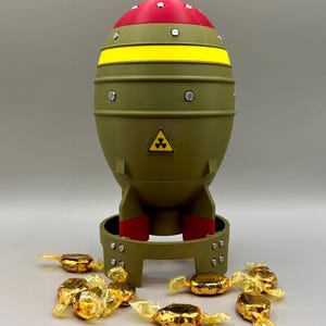 Peut inclure: Un distributeur de bonbons fantaisie en forme de bombe, avec un dessus rouge, une bande jaune et un corps vert olive. Le distributeur porte un symbole de radiation et est monté sur un support. Des bonbons dorés sont éparpillés autour de la base.