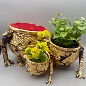 Op de afbeelding: Drie gestructureerde goudkleurige en zwarte potten met pootjes, gegroepeerd opgesteld. De grootste pot heeft een rode fluwelen bovenkant. De twee kleinere potten bevatten kunstmatige groene planten met kleine gele bloemen. Verweerde afwerking.