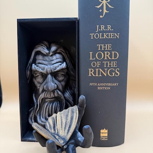 May include: A black and gray sculpture of a bearded man in a black box, with a hand holding a butterfly. Next to it is a dark blue book with gold lettering that reads "The Lord of the Rings" by J.R.R. Tolkien, 50th Anniversary Edition.