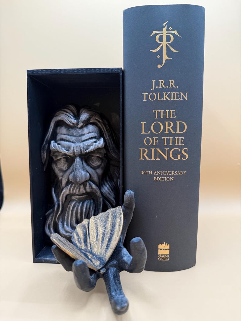 May include: A black and gray sculpture of a bearded man in a black box, with a hand holding a butterfly. Next to it is a dark blue book with gold lettering that reads "The Lord of the Rings" by J.R.R. Tolkien, 50th Anniversary Edition.