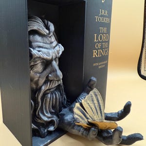 May include: A black bookend featuring a detailed sculpture of a bearded figure's head and hand. The hand holds a gold-colored object. The bookend is next to a copy of "The Lord of the Rings" book.