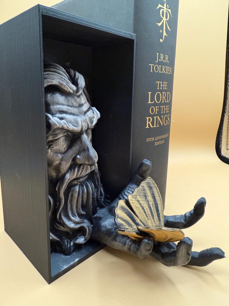 May include: A black bookend featuring a detailed sculpture of a bearded figure's head and hand. The hand holds a gold-colored object. The bookend is next to a copy of "The Lord of the Rings" book.
