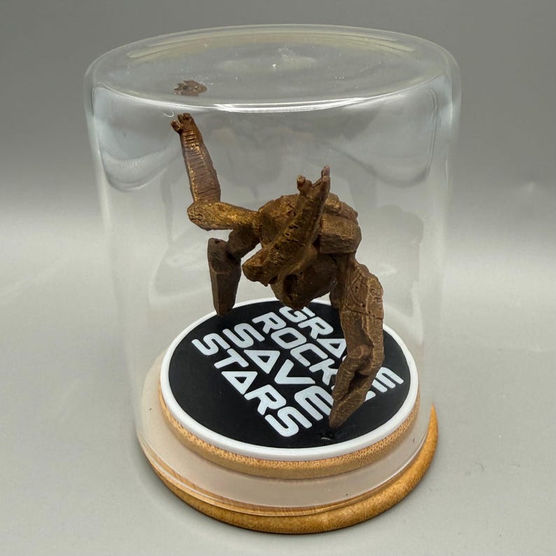May include: A bronze-colored, textured rock-like figure stands inside a clear glass dome on a circular base. The base features a black circular insert with the white text "GRAVITY ROCKS SAVE STARS" printed in a bold, sans-serif font.
