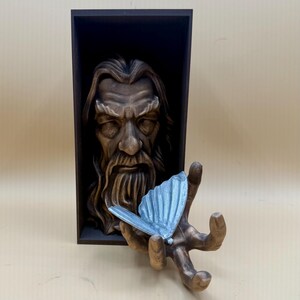 May include: A brown sculpture of a wise man's face in a dark wooden frame. A hand, also brown, holds a light blue butterfly. The sculpture has a detailed beard and long hair. The butterfly has detailed wings.