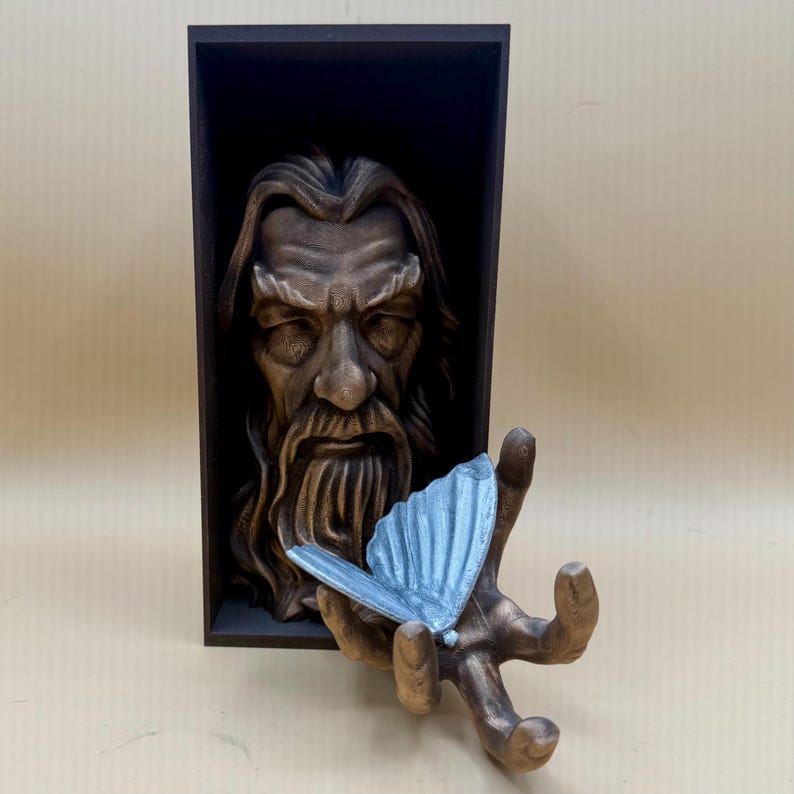 May include: A brown sculpture of a wise man's face in a dark wooden frame. A hand, also brown, holds a light blue butterfly. The sculpture has a detailed beard and long hair. The butterfly has detailed wings.