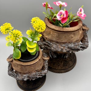 Op de afbeelding: Twee bronskleurige plantenbakken met gestructureerde wervelpatronen en ruwe, rotsachtige voetstukken. De kleinere bak bevat gele bloemen met groene bladeren, terwijl de grotere bak roze en witte bloemen met groen blad bevat.
