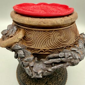 Puede incluir: Un recipiente marrón en forma de vasija con intrincados patrones de remolinos en el cuerpo y dos esculturas plateadas y texturizadas en forma de manos que sujetan los lados. La parte superior tiene una tapa circular de color rojo brillante.