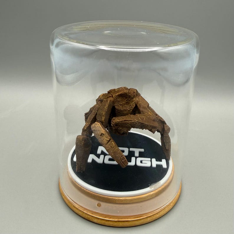 May include: A brown, textured, multi-legged creature figurine enclosed in a clear glass dome. The figure rests on a circular black base with the white text "NOT ENOUGH" printed on it. The base features a light-colored wooden rim at the bottom.