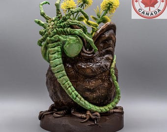 Puede incluir: Una maceta marrón con textura en forma de huevo que presenta una criatura verde de múltiples patas sobre el borde. La maceta contiene varias flores artificiales amarillas. Un logotipo circular en la esquina superior derecha dice Made in Canada con una hoja de arce.