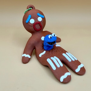 May include: A brown gingerbread man figurine with white icing details and a blue monster emerging from its chest. The gingerbread man has a red mouth, blue teardrops, and a green beaded loop at the top.