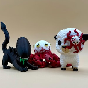 以下が含まれることがあります： 3Dプリントされた3つのフィギュア：緑色のよだれを垂らした黒いエイリアン風の生き物、赤と白のタコ、顔に赤いタコが付いた白い羊。 フィギュアはベージュ色の背景にあります。