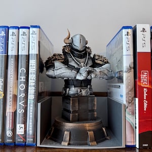 May include: A silver and bronze bust of a character with horns, a helmet, and armour, positioned between video game cases. The bust is on a tiered base. Video game cases include "Metal Gear Solid," "Chorvs," and "Death Stranding."