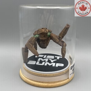 May include: A brown, textured, multi-limbed creature figurine with green accents, enclosed in a clear glass dome on a circular wooden base. The base features a black circular insert with the white text FIST MY BUMP printed in bold letters.
