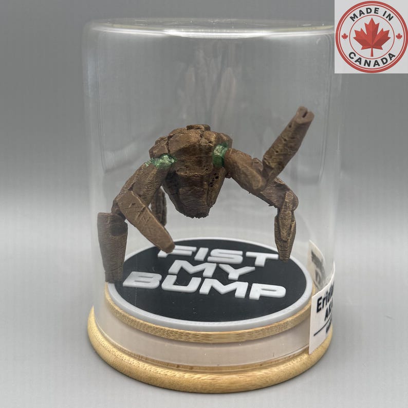 May include: A brown, textured, multi-limbed creature figurine with green accents, enclosed in a clear glass dome on a circular wooden base. The base features a black circular insert with the white text FIST MY BUMP printed in bold letters.
