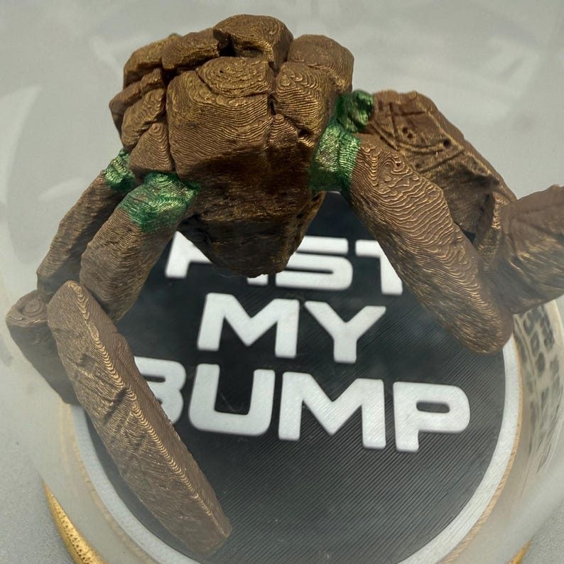 May include: A 3D printed brown rock-like creature figure with green accents positioned over a circular black base. The base features the white text FIST MY BUMP in bold capital letters. The figure has a textured, layered surface appearance.