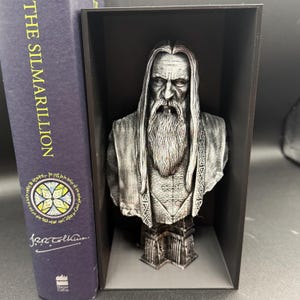 May include: A silver-colored bust of a bearded man with long hair, displayed in a black box. The bust is next to a book titled "The Silmarillion" with a circular design on the cover. The bust has intricate details.