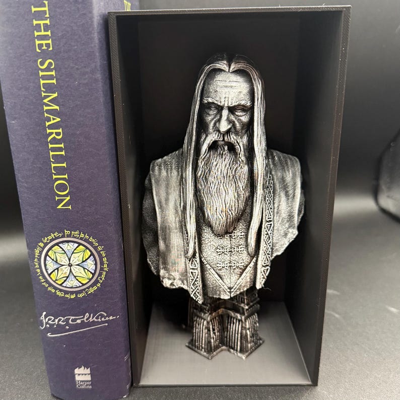May include: A silver-colored bust of a bearded man with long hair, displayed in a black box. The bust is next to a book titled "The Silmarillion" with a circular design on the cover. The bust has intricate details.