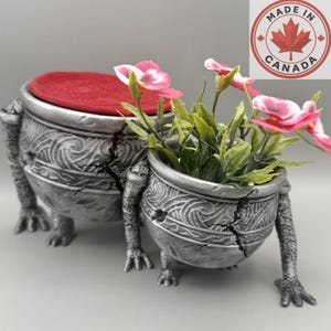 May include: Two silver-toned, textured, cauldron-shaped planters with three clawed feet and intricate swirling patterns. The larger planter has a red circular lid, while the smaller one contains artificial pink flowers with green leaves.