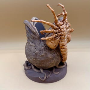 May include: A detailed sculpture of a creature emerging from an egg. The egg is grey with a textured surface, and the creature is brown with multiple limbs. The sculpture is on a grey base.