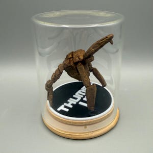 May include: A brown, multi-jointed, articulated figure stands on a circular black base with the white text THUMB IT printed on it. The figure is enclosed within a clear, cylindrical glass display dome with a light-colored wooden base.
