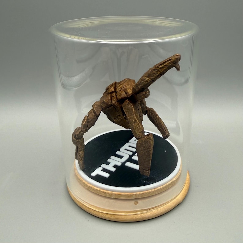May include: A brown, multi-jointed, articulated figure stands on a circular black base with the white text THUMB IT printed on it. The figure is enclosed within a clear, cylindrical glass display dome with a light-colored wooden base.