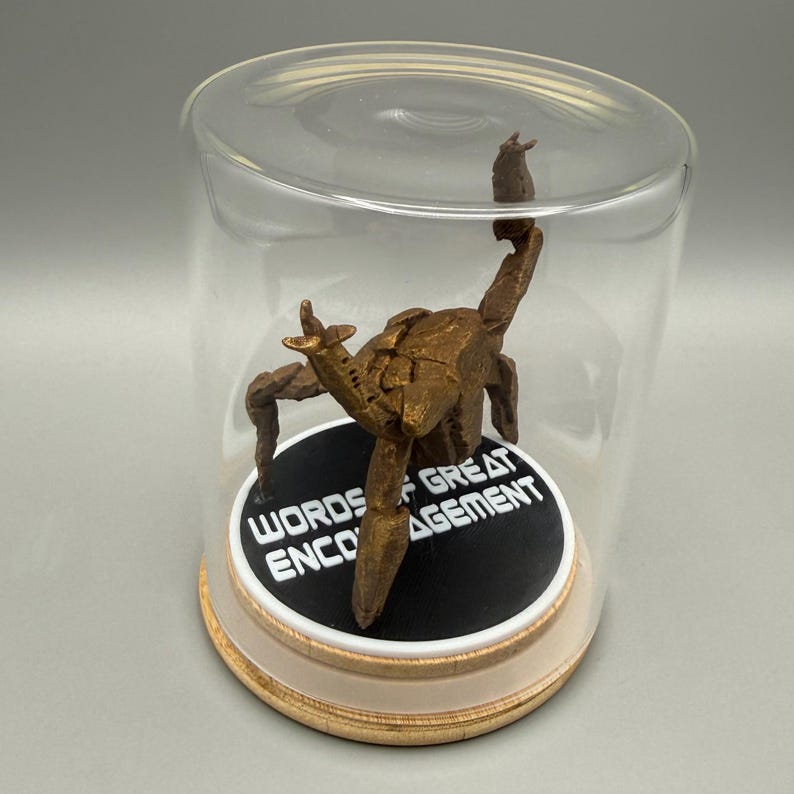 May include: A bronze-colored, multi-legged creature figurine enclosed in a clear glass dome on a light wood base. The creature stands on a black circular platform featuring the white text "WORDS OF GREAT ENCOURAGEMENT" printed in a bold, sans-serif font.