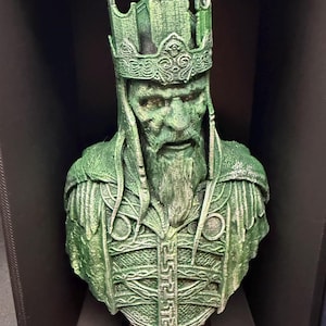 May include: A green bust of a king with a detailed crown and beard. The sculpture has intricate details on the armor and face, with a weathered appearance. The bust is displayed on a dark surface.