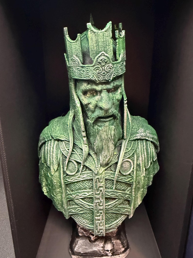 May include: A green bust of a king with a detailed crown and beard. The sculpture has intricate details on the armor and face, with a weathered appearance. The bust is displayed on a dark surface.