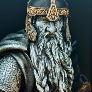 May include: A detailed bust of a dwarf warrior with a long, braided beard and a helmet. The helmet and decorative trim are gold-colored, while the face and beard are a lighter gray. The warrior's armor is visible on the shoulders.